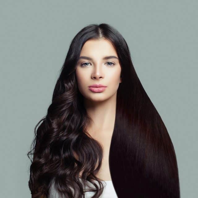 Zdjęcie przedstawiające kobietę stojącą przodem w planie amerykańskim z włosami w kolorze ciemnego brązu. Włosy z prawej strony są gładkie, lśniące i proste, natomiast włosy z lewej strony są pokręcone. W ten sposób przedstawiono, jak wyglądają proste włosy po zabiegu nanoplastii.