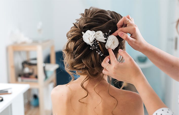 Zdjęcie przedstawiające Pannę Młoda podczas wykonywania fryzury w dniu ślubu. Na zdjęci widoczne jest upięcie ślubne z tyłu oraz dłonie fryzjera stylisty wpinające we włosy ślubne ozdoby. 