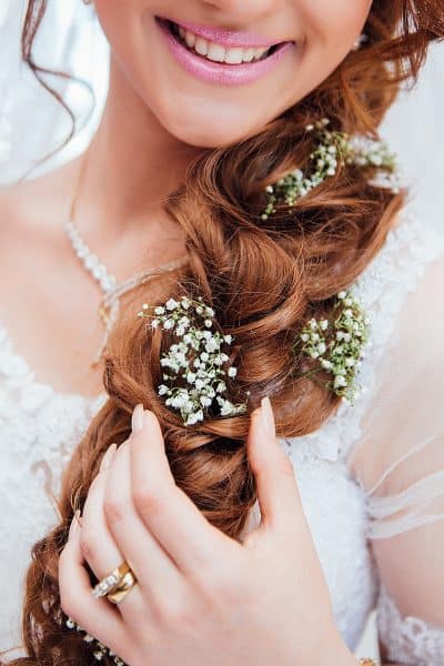 Zdjęcie przedstawiające upięcie ślubne w postaci luźno spleconego warkocza opadającego na przód. Włosy w kolorze kasztanowym ozdobione kwiatami białej gipsówki. Ujęcie portretowe przodem z fokusem na włosy, bez widocznej twarzy. 