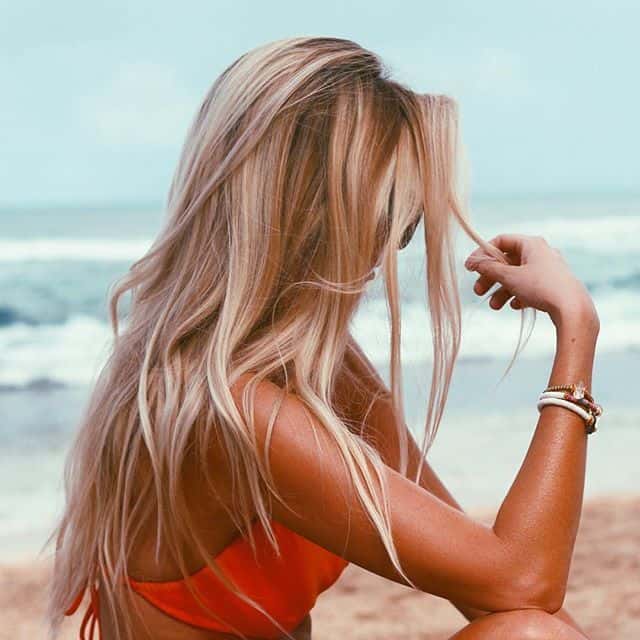 Na zdjęciu widoczna kobieta z długimi, rozpuszczonymi i potarganymi wiatrem włosami koloru blond siedząca na plaży na tle morza. Kadr amerykański bokiem. 
