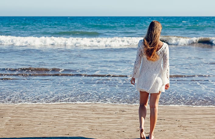  Na zdjęciu widoczna kobieta z rozpuszczonymi, długimi blond włosami stojąca tyłem na plaży. Patrzy w kierunku morza. Kadr szeroki. 
