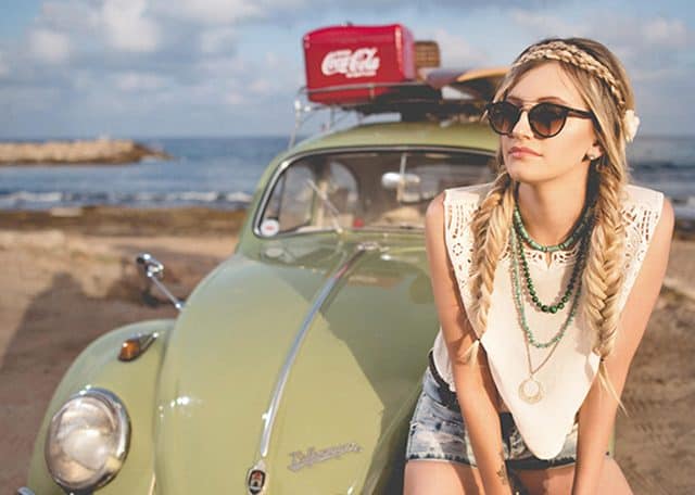 Na zdjęciu widoczna dziewczyna o blond włosach splecionych w warkocze siedząca na masce samochodu Volkswagen Garbus w kolorze groszkowym. W tle widoczna plaża oraz może. Kadr amerykański.