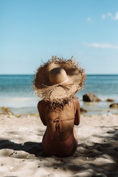 Zdjęcie przedstawiające kobietę w kostiumie kąpielowym siedzącą na plaży na tle morza oraz nieba. Jej włosy schowane są pod słomianym kapeluszem. Ujęcie z tyłu. 