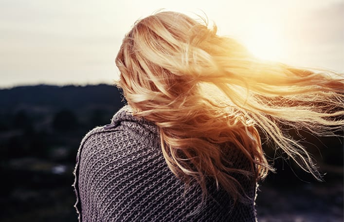  Na zdjęciu widoczna kobieta stojąca tyłem z długimi blond włosami rozwianymi przez wiatr i podświetlonymi słońcem. Kadr amerykański.