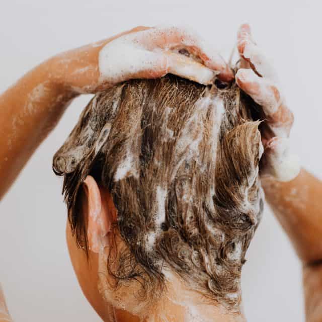 Na zdjęciu widoczna kobieta z długimi, mokrymi blond włosami. Jej dłonie dotykają czubka głowy, aby zwrócić uwagę na skórę głowy. Kadr portretowy. 