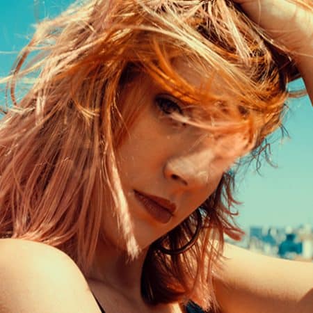Na zdjęciu widoczna kobieta z rozwianymi wiatrem i oświetlonymi słońcem rudymi włosami średniej długości. Kadr portretowy.