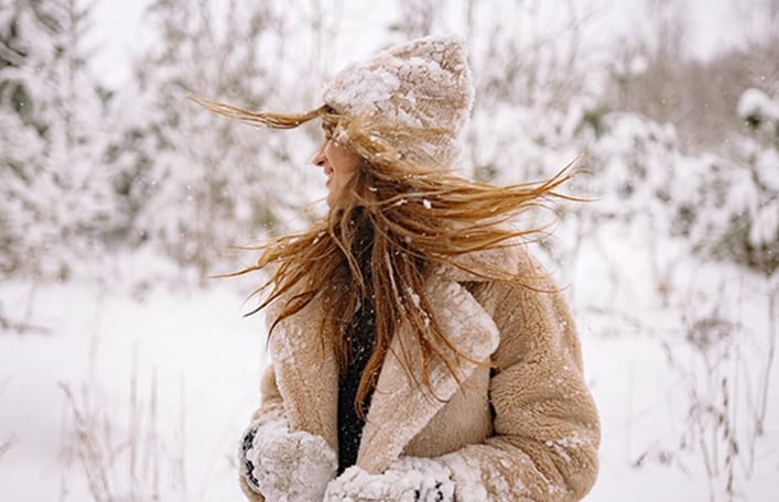 Na zdjęciu widoczna kobieta z rozwianymi długimi włosami w kolorze ciemnego blondu. Kobieta ubrana jest w czapkę oraz grubą kurtkę. Sceneria zimowa. Kadr amerykański. 