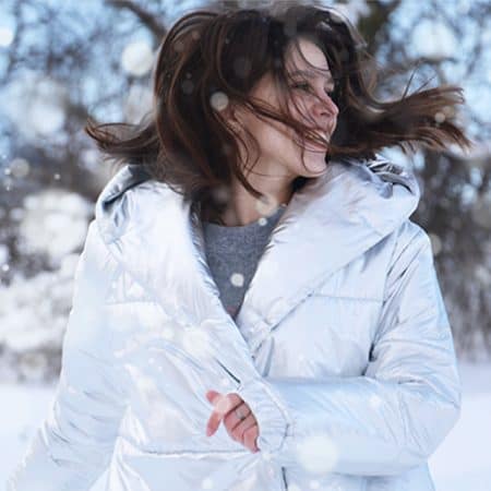 Na zdjęciu widoczna roześmiana kobieta z ciemnymi włosami potarganymi przez wiatr. Sceneria zimowa. Kadr amerykański.