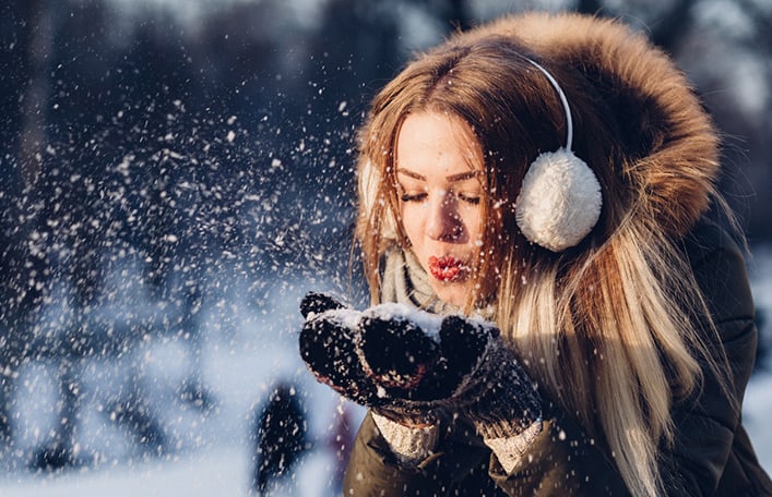 Na zdjęciu widoczna kobieta ubrana w nauszniki i zimowa kurtkę z rozpuszczonymi, długimi włosami, która bawi się śniegiem. Sceneria zimowa. Kadr amerykański. 