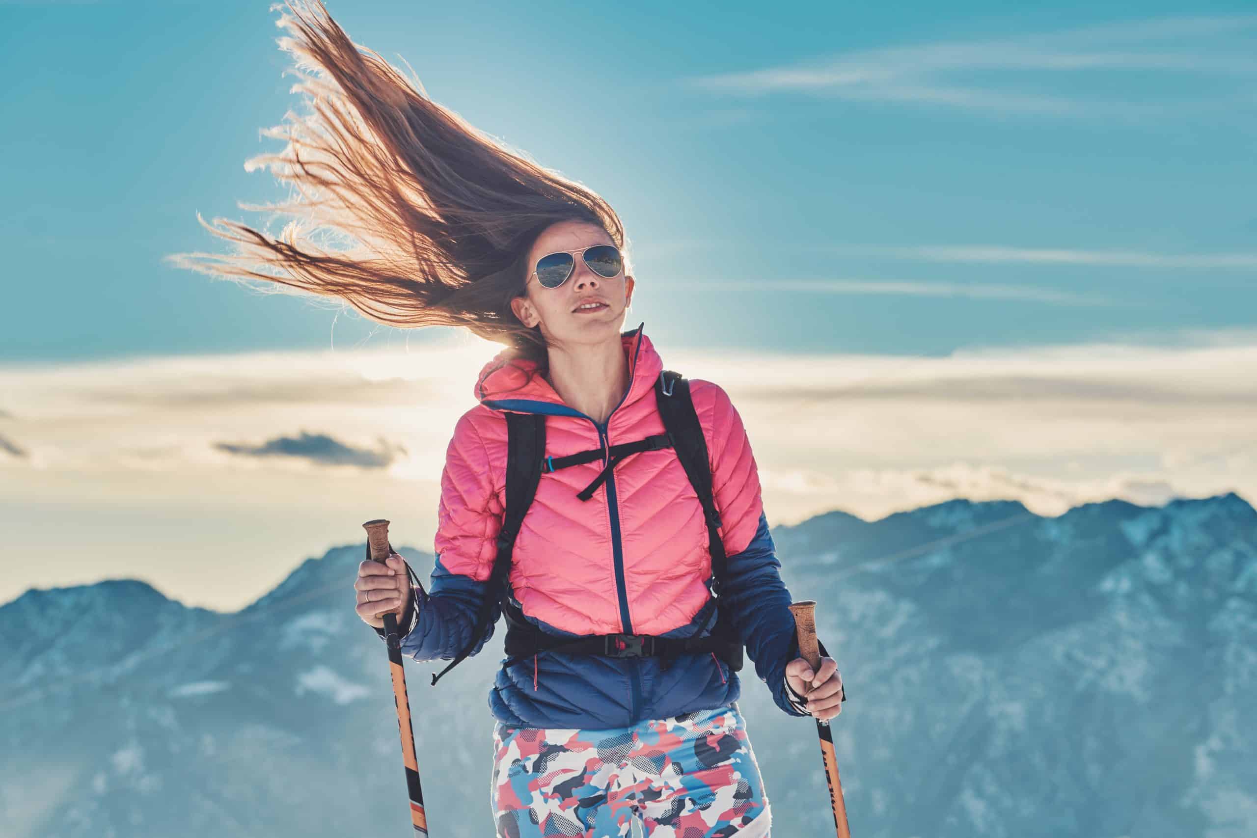 Na zdjęciu widoczna kobieta jeżdżąca na nartach, z długimi mocno rozwianymi przez wiatr włosami. Sceneria zimowa. Kadr amerykański. 