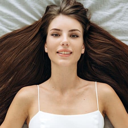 Na zdjęciu widoczna kobieta leżąca na łóżku, z bardzo gęstymi i długimi włosami w kolorze brązu. Włosy są rozłożone, co optycznie jeszcze bardziej zwiększa ich objętość. Ujęcie od góry.