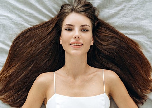 Na zdjęciu widoczna kobieta leżąca na łóżku, z bardzo gęstymi i długimi włosami w kolorze brązu. Włosy są rozłożone, co optycznie jeszcze bardziej zwiększa ich objętość. Ujęcie od góry.