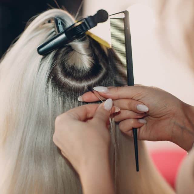 Na zdjęciu widoczny tył kobiecej głowy, z podzielonymi na sekcje i podpiętymi blond włosami, podczas zabiegu przedłużania włosów metodą keratynową.