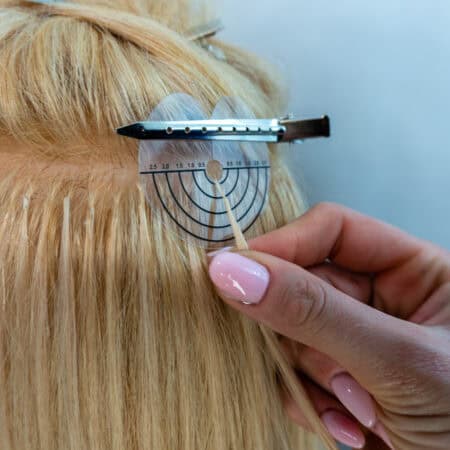 Na zdjęciu widoczny tył kobiecej głowy, z podzielonymi na sekcje i podpiętymi blond włosami, podczas zabiegu przedłużania włosów metodą keratynową.