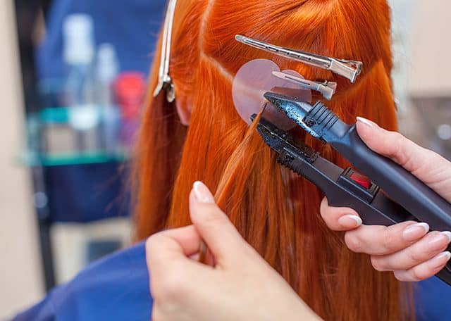 Na zdjęciu widoczny tył kobiecej głowy z rudymi włosami, podczas zabiegu przedłużania włosów metodą keratynową.