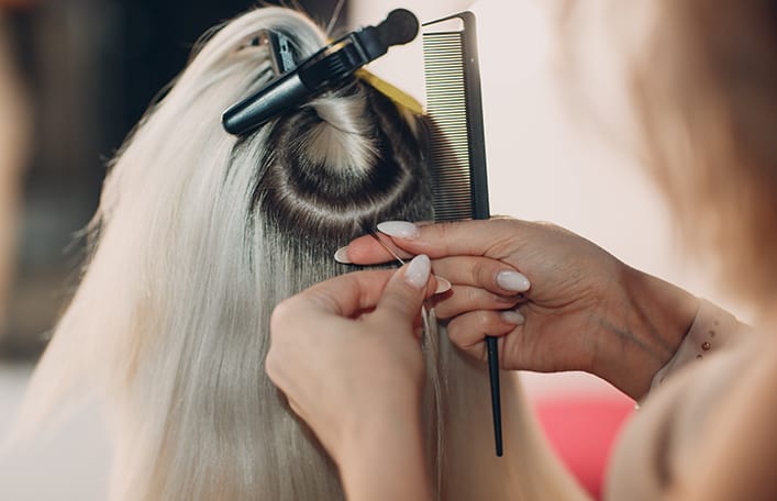 Na zdjęciu widoczny jest tył kobiecej głowy z podpiętymi pasmami blond włosów, podczas tworzenia mini kosmyków w trakcie zabiegu przedłużania włosów metodą keratynową. 