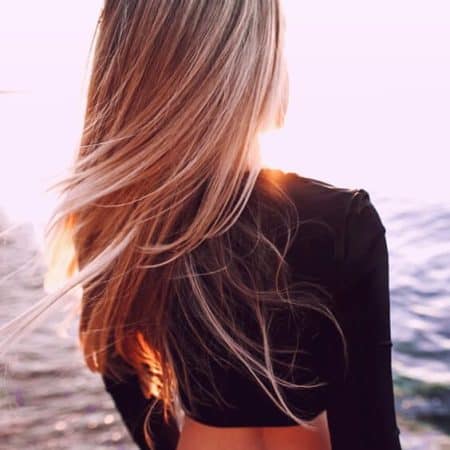 Na zdjęciu widoczny tył kobiecej sylwetki z rozwianymi przez wiatr długimi włosami. Kobieta stoi na plaży na tle zachodzącego słońca.