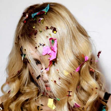 Na zdjęciu widać kobiecą głowę z blond włosami opadającymi na twarz oraz z lecącym z góry kolorowym konfetti.