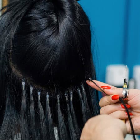 Zdjęcie przedstawiam kobietę z czarnymi włosami tyłem, u której wykonywane jest przedłużanie włosów metodą na nano ringi.